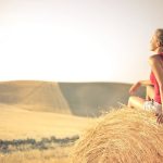 Woman with a backpack sitting on a hay bale, enjoying a serene sunset over a vast countryside field.