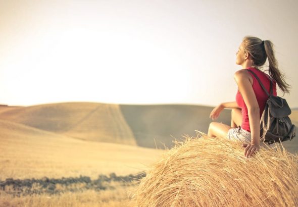 Woman with a backpack sitting on a hay bale, enjoying a serene sunset over a vast countryside field.