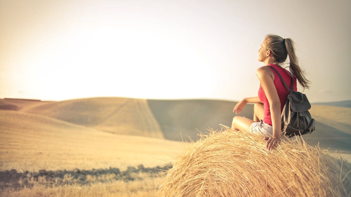 Woman with a backpack sitting on a hay bale, enjoying a serene sunset over a vast countryside field.
