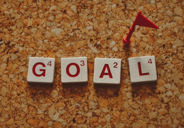 Scrabble tiles forming the word 'goal' with a small red flag on a cork board background.